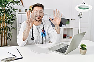 Young doctor working at the clinic using computer laptop showing and pointing up with fingers number eight while smiling confident