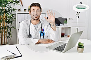 Young doctor working at the clinic using computer laptop showing and pointing up with fingers number five while smiling confident