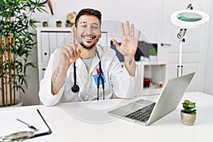Young doctor working at the clinic using computer laptop showing and pointing up with fingers number six while smiling confident