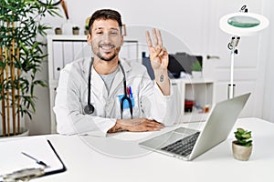 Young doctor working at the clinic using computer laptop showing and pointing up with fingers number three while smiling confident