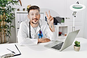 Young doctor working at the clinic using computer laptop showing and pointing up with fingers number two while smiling confident