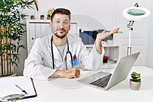 Young doctor working at the clinic using computer laptop smiling cheerful presenting and pointing with palm of hand looking at the