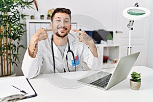 Young doctor working at the clinic using computer laptop smiling cheerful showing and pointing with fingers teeth and mouth