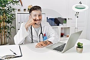 Young doctor working at the clinic using computer laptop smiling doing phone gesture with hand and fingers like talking on the