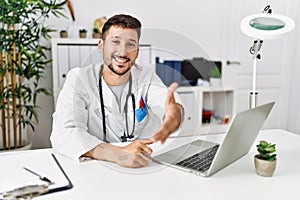 Young doctor working at the clinic using computer laptop smiling friendly offering handshake as greeting and welcoming