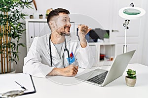 Young doctor working at the clinic using computer laptop smiling with happy face looking and pointing to the side with thumb up