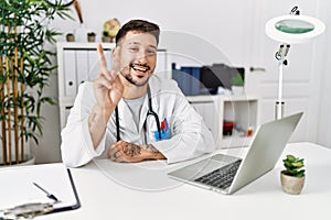 Young doctor working at the clinic using computer laptop smiling with happy face winking at the camera doing victory sign