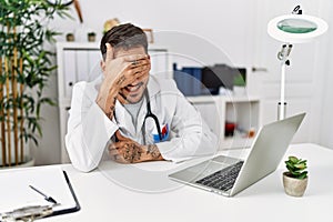 Young doctor working at the clinic using computer laptop smiling and laughing with hand on face covering eyes for surprise