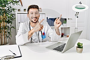 Young doctor working at the clinic using computer laptop smiling and looking at the camera pointing with two hands and fingers to