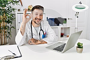 Young doctor working at the clinic using computer laptop smiling positive doing ok sign with hand and fingers