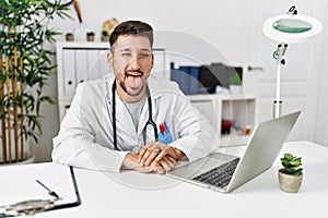 Young doctor working at the clinic using computer laptop sticking tongue out happy with funny expression