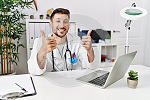 Young doctor working at the clinic using computer laptop success sign doing positive gesture with hand, thumbs up smiling and