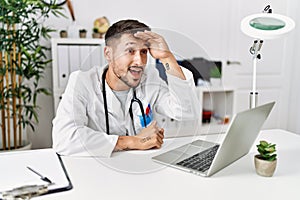 Young doctor working at the clinic using computer laptop very happy and smiling looking far away with hand over head