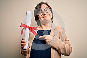 Young down syndrome business woman holding univeristy diploma award over isolated background very happy pointing with hand and photo