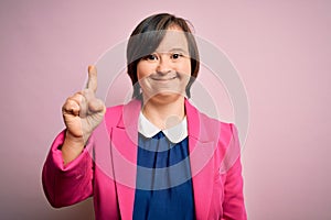 Young down syndrome business woman over pink background showing and pointing up with finger number one while smiling confident and