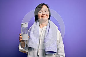Young down syndrome fitness woman training at gym holding water bottle and sport towel with a happy face standing and smiling with