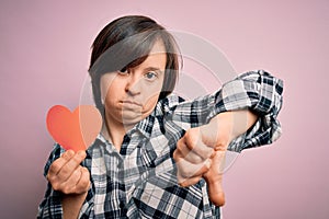 Young down syndrome romantic woman holding red heart paper shape over pink background with angry face, negative sign showing