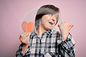 Young down syndrome romantic woman holding red heart paper shape over pink background pointing and showing with thumb up to the