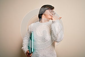 Young down syndrome student woman reading a book from library over isolated background pointing and showing with thumb up to the