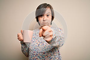 Young down syndrome woman drinking a cup of hot coffee over isolated background pointing with finger to the camera and to you,