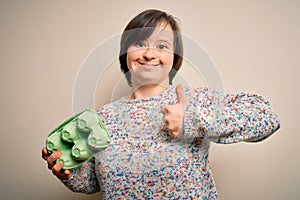 Young down syndrome woman holding cardboard egg cup from fresh healthy eggs happy with big smile doing ok sign, thumb up with