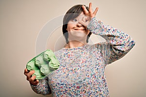 Young down syndrome woman holding cardboard egg cup from fresh healthy eggs with happy face smiling doing ok sign with hand on eye