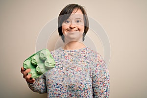 Young down syndrome woman holding cardboard egg cup from fresh healthy eggs with a happy face standing and smiling with a