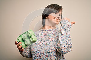 Young down syndrome woman holding cardboard egg cup from fresh healthy eggs pointing and showing with thumb up to the side with