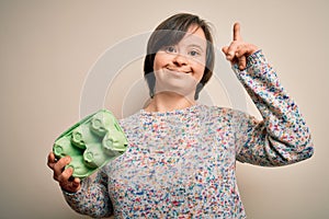 Young down syndrome woman holding cardboard egg cup from fresh healthy eggs surprised with an idea or question pointing finger