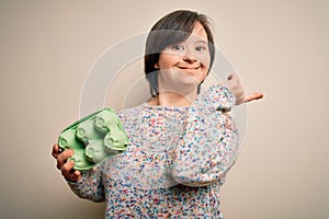 Young down syndrome woman holding cardboard egg cup from fresh healthy eggs very happy pointing with hand and finger to the side