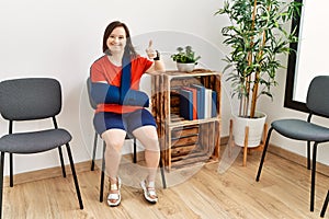 Young down syndrome woman sitting at doctor waiting room with arm injury doing happy thumbs up gesture with hand