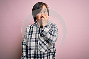 Young down syndrome woman wearing casual shirt over pink background looking stressed and nervous with hands on mouth biting nails