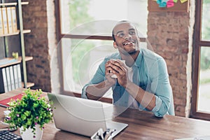 Young dreaming mulatto american worker is thinking in front of laptop at work place. He is happy, smiling, behind him is window an