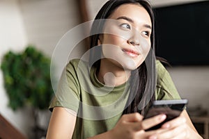 Young dreamy woman looking outside window, holding smartphone and smiling, chatting on phone app