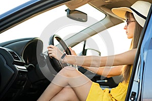 Young driver girl in yellow dress and straw hat sitting behind steering wheel driving a car. Summer vacation and travel concept