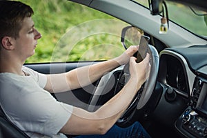 Young driver guy distracted by his phone while in front of the steering wheel, using his smartphone with one hand while driving.