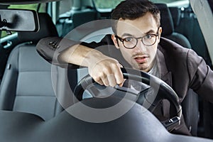 Young driver is looking at the camera while driving his car. He holding steering wheel with both hands