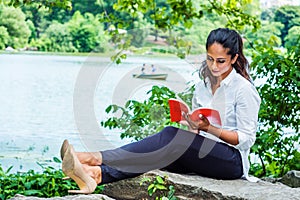 Young East Indian American Woman reading red book, relaxing at Central Park, New York