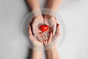 Young and elderly women holding red heart on light grey background, top view