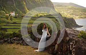 Young elegant bride in white long wedding dress. Romantic background of beautiful idyllic green valley and ocean coast