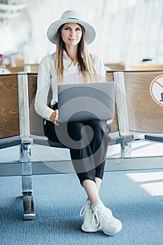 Young elegant business woman with hand luggage in international airport terminal, working on her laptop while waiting