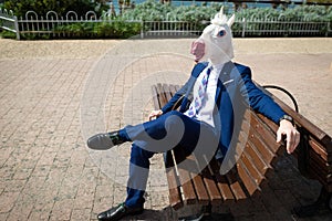 Young elegant man in funny mask and suit have a break in park