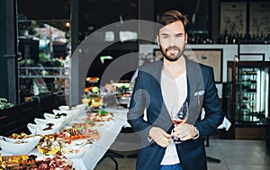 Young elegant man standing in the restaurant, holding a glass of wine. Man's style