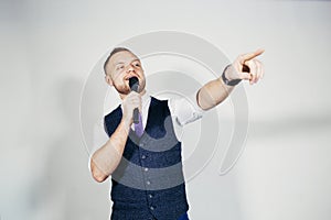 Young elegant talking man holding microphone talking with pointing finger. Isolated on grey background.