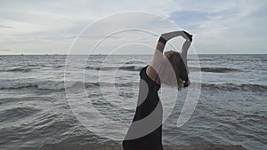 Young elegant woman in black long dress dancing with hands up holding black scarf on the pier