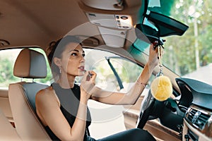 Young elegant woman looking in the car view mirror while applying makeup, lipstick on the lips