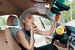 Young elegant woman looking in the car view mirror while applying makeup, lipstick on the lips