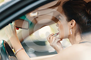 Young elegant woman looking in the car view mirror while applying makeup, lipstick on the lips