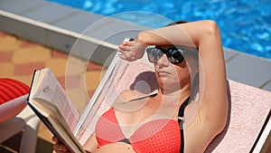 Young, elegant woman reading book on sunbed by pool