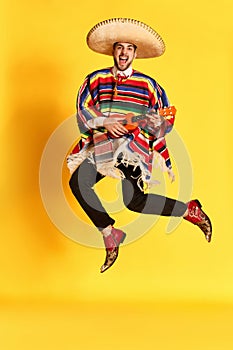 Young emotional man in colorful poncho and sombrero posing, playing guitar and jumping against yellow studio background